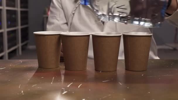Рука чоловіка в білій лабораторній шубці наповнює воду з пластикової пляшки до чотирьох паперових чашок
 - Кадри, відео