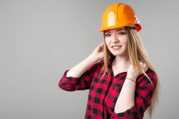 Portrait of a young attractive woman with blond hair in orange helmet on a neutral gray background. - Foto, Imagen