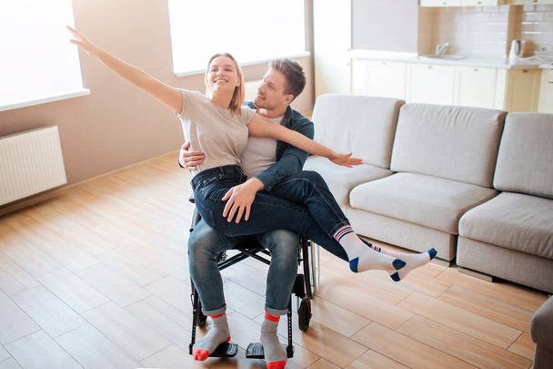 Young man with inclusiveness holding girlfirend on knees. She is happy and full of joy. They smile. Person with special needs. Together in empty room. - 写真・画像