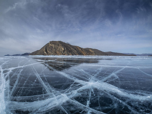 Прозорий лід з тріщинами і рифтами на озері Байкал біля замороженого острова з камінням, покритими снігом і Хоар Морозом. Гладка слизька поверхня виглядає як скло з відображенням під небом з хмарами. - Фото, зображення