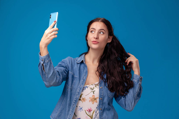 Молода брюнетка позує зі смартфоном, що стоїть на синьому фоні
. - Фото, зображення