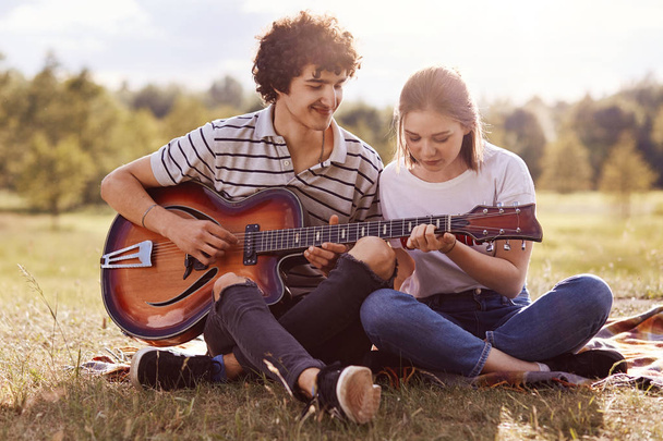 Image of young handsome man with dark curly hair teaches playing guitar her friend, couple have picnic in meadow, have romantic, enjoying spending time together, hot sunny summer day. Youth concept. - Fotó, kép