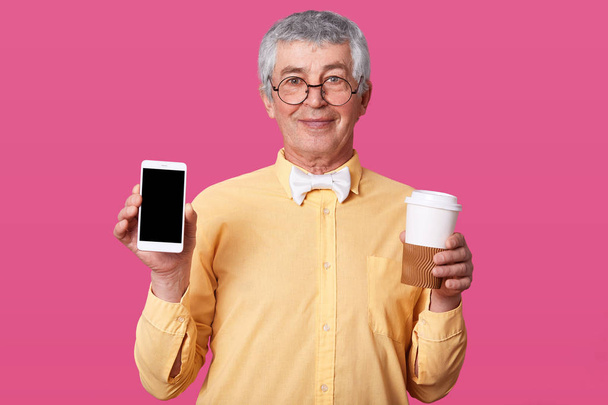 Close up portrait of mature grey haired man in yellow shirt with bow tie, holds mobile phone with blank screen and takeaway coffee, looks directly at camera, isolated over pink studio background. - Foto, Imagen
