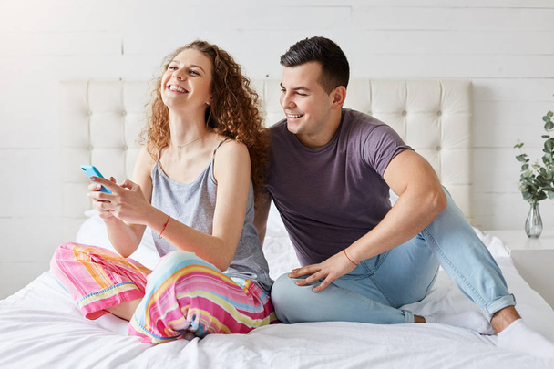 Happy couple spend morning time in bedroom, checking social network via smartphone, being always in touch, communicate with their friends, Family with mobile phones sitting laughing in comfortable bed - Photo, Image