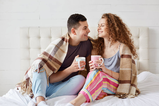 Happy couple relaxing in bed with hot beverage, holds cups full of tea or coffee, sit under warm blanket, dresses casually, romantic young family, spend spare time together at home. Relation concept. - Foto, imagen