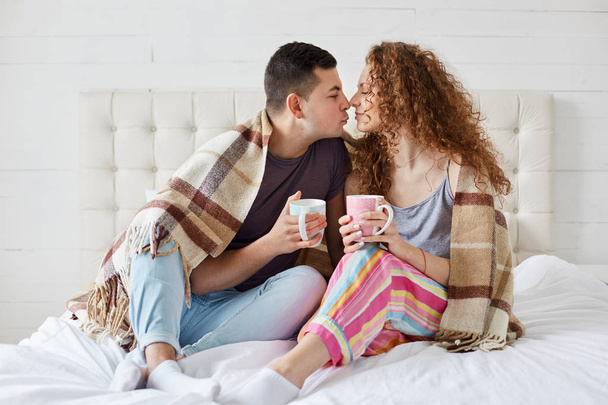 Indoor shot of happy female and male have romantic relationships, drink coffee or tea, covered with plaid in bedroom. Romantic couple enjoying spending leisure together. Relationship concept. - Foto, Imagen
