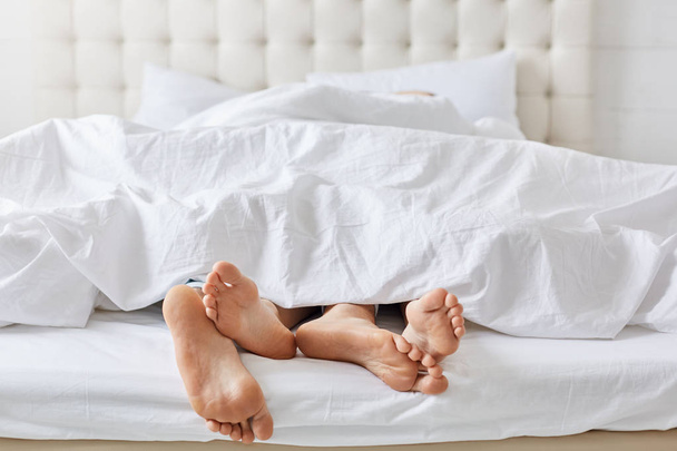 Horizontal shot of feet of couple under white bedclothes in bed at bedroom, enjoying good rest at home and calm atmosphere. Female and male relaxing together at weekend. Pomantic couple in love. - Foto, Imagem