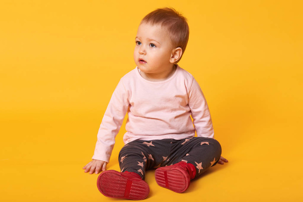 Studio shot of adorable little baby girl sitting on floor, isolated on yellow background, lovely baby portrait wearing casual clothes and red shoes, looks aside, looking for her mother. Copy space. - Zdjęcie, obraz