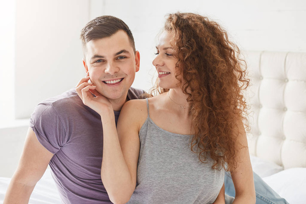 Woman in pyjamas sits with her husband early in morning, sits on bed, touches him gently with hand. Pretty curly female spend time with boyfriend. Handsome male looks smiling at camera. Happy couple. - Foto, immagini