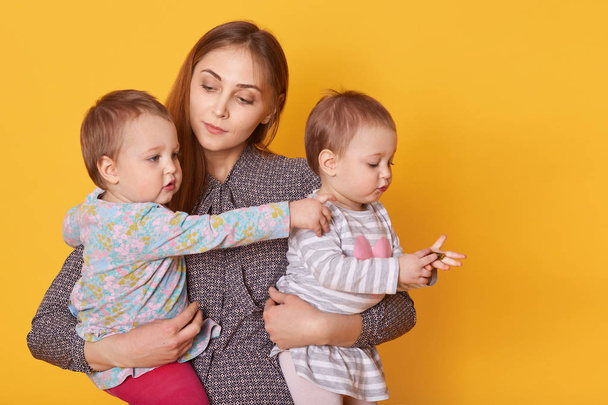Portrait of responsible attentive mother spending time with her little children, holding twin girls in arms, listening to them seriously. Cute kid rises her arm, showing direction, looks curious. - Foto, Bild