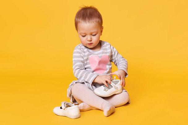 Fair haired pretty little child wearing stripped dress with heart poses isolated over bright yellow background. Sweet magnetic kid sits on floor in studio holding and observing her white shoes. - Valokuva, kuva