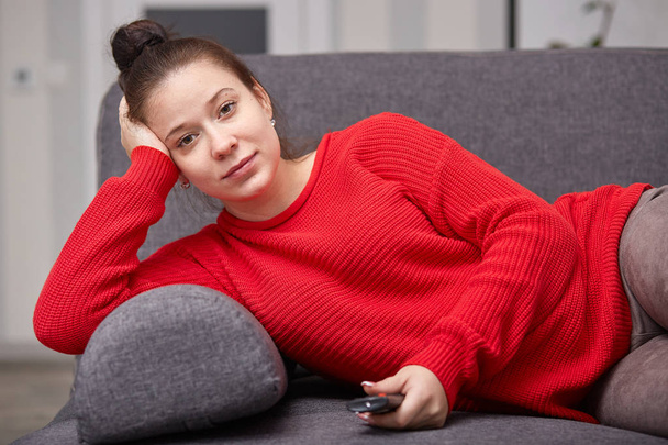 Shot of pregnant female watches TV at home, holds remote control and switches channels, enjoying interesting TVshow, lies on gray sofa, looking at monitor. Pregnancy, relax and free time concept. - Foto, Imagen