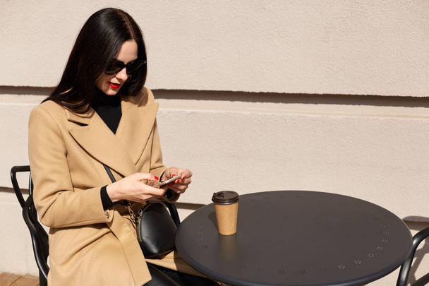 Close up portrait of beautiful elegant young woman wearing beige coat and drinking takeaway coffee while having lunch time. Pretty brunette lady checking her social networking sites, chatting. - Φωτογραφία, εικόνα