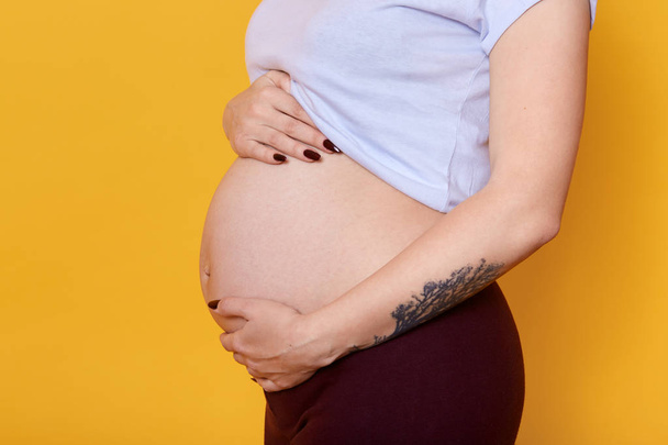 Side view of pregnant female with naked belly isolated over yellow background. Woman with tattoo on hend dressed casually being photographed in photo studio. Motherhood and pragnancy concept. - Photo, Image