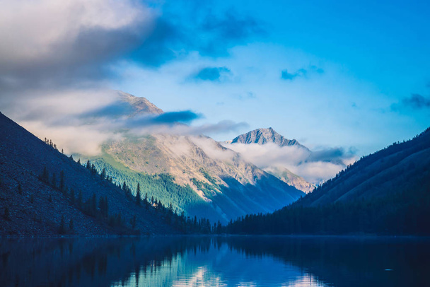 Amazing blue silhouettes of mountains under blue cloudy sky. Beautiful ripples on water of mountain lake. Low clouds before mountain ridge. Wonderful highland landscape. Picturesque mountainscape. - Photo, Image