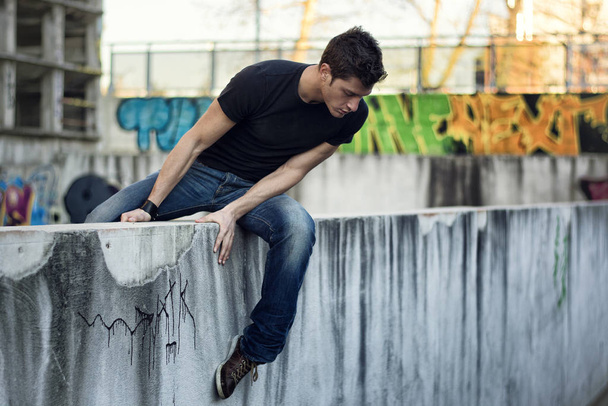 Young man sitting and balancing on dirty wall, looking down, urban environment - Fotoğraf, Görsel