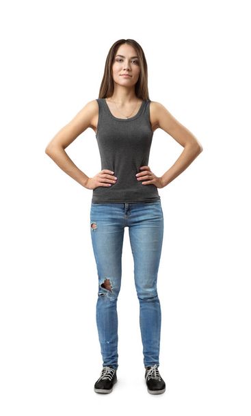Front view of young attractive woman in gray sleeveless top and blue jeans standing with arms on waist isolated on white background. - Foto, afbeelding