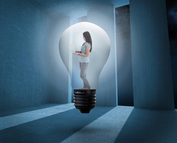 Woman holding laptop in light bulb against light shining into dark room - Фото, изображение