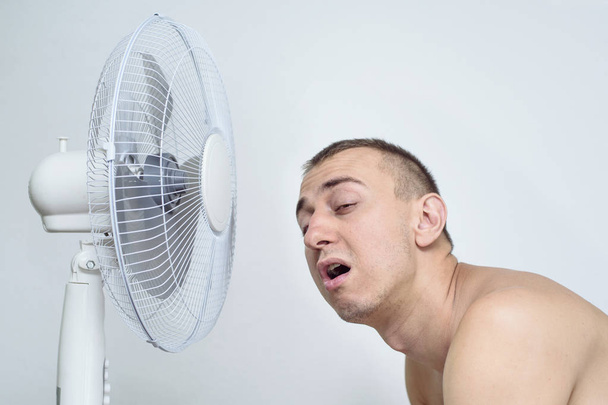 Hombre con rastrojo en su cara sufre del calor y tratando de refrescarse cerca del ventilador
. - Foto, Imagen