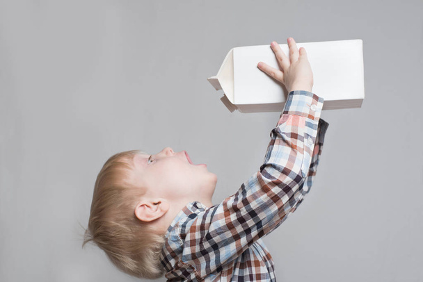 Little blond boy drinks from a large white package. Light background - Foto, imagen