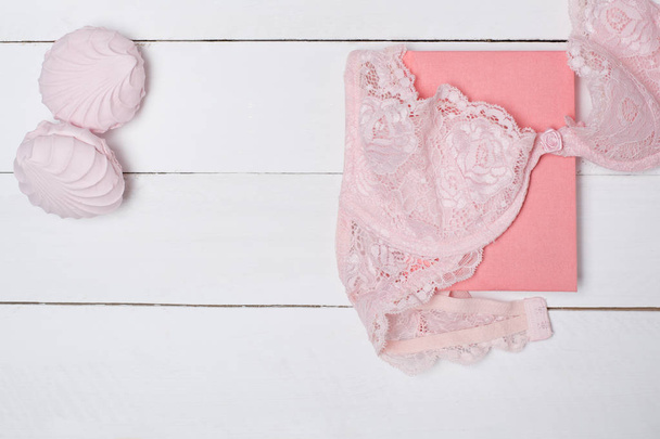Corpiño de encaje rosa, bloc de notas y malvavisco sobre fondo de madera blanca. Concepto de moda
. - Foto, Imagen