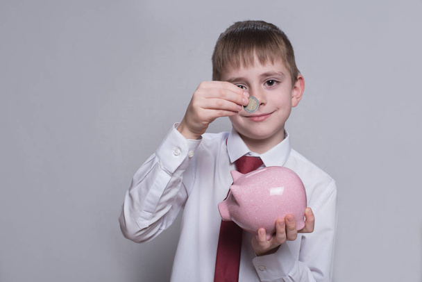 Το χαμογελαστή αγόρι βάζει ένα κέρμα σε ένα ροζ κουμπαράς. Επιχειρηματική ιδέα. Φως φόντο - Φωτογραφία, εικόνα