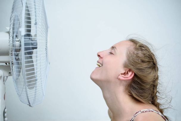 Jeune femme assise sous un ventilateur avec les yeux fermés du plaisir. Chaleur estivale. Fond blanc
 - Photo, image