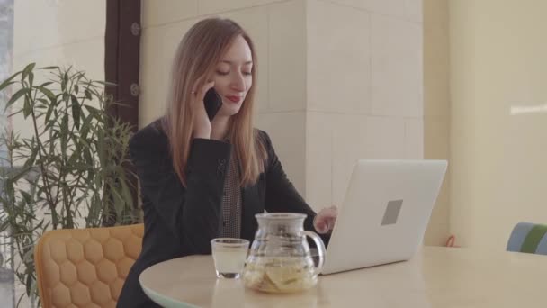 Adult woman is talking by mobile phone and typing on notebook in her office - Imágenes, Vídeo