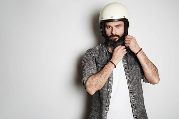 Brutal bearded hipster male model poses in black jeans shirt and blank white t-shirt premium summer cotton with moto helmet in hands, on white background. Copy paste Advertisement. - Foto, Imagem