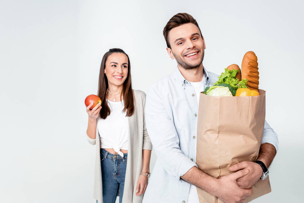 Happy young woman with apple looking at smiling man holding grocery bag - Foto, imagen
