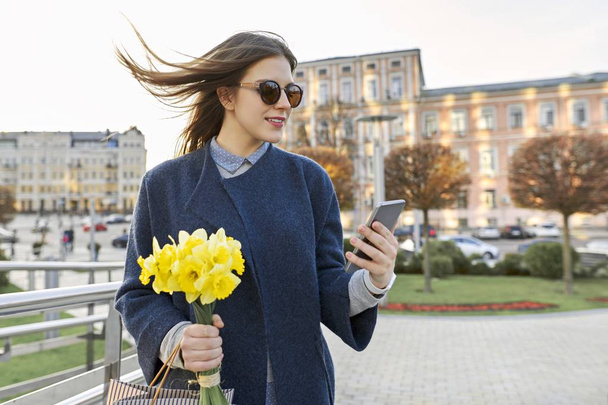 Beautiful girl with bouquet of yellow spring flowers, young woman reading text with smartphone, background city architecture, golden hour - Photo, Image