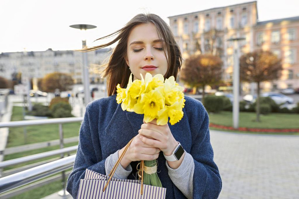 Portrait of young woman with bouquet of yellow spring flowers daffodils. Beautiful girl in city enjoys flowers, eyes closed. - Foto, imagen