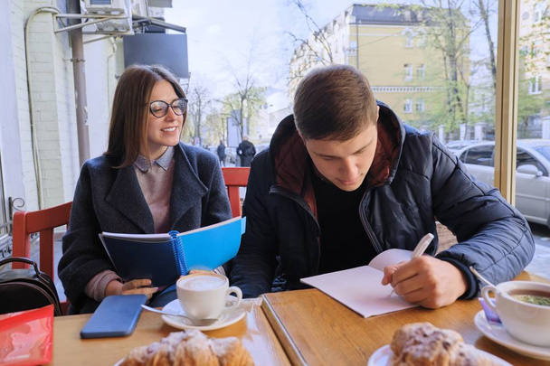 Young couple of students study in outdoor cafe, drink coffee tea, eat croissants, background is spring city street - 写真・画像