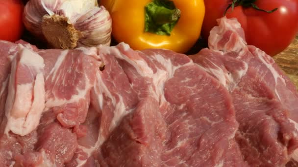 Raw meat steaks are cut from a large piece of pork or beef on a wooden cutting board. Vegetables: tomatoes, sweet peppers, garlic. Close-up. - Footage, Video