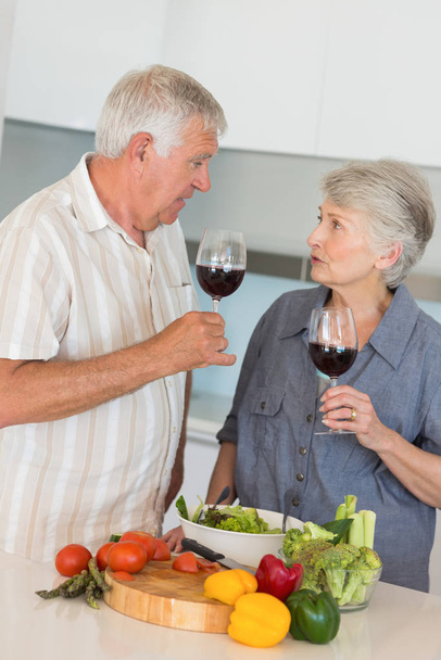 Sonriendo pareja mayor preparando una ensalada y teniendo vino tinto en casa en la cocina
 - Foto, imagen