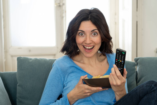 Lifestyle portrait of cheerful young woman sitting on the couch watching TV holding remote control - Foto, afbeelding