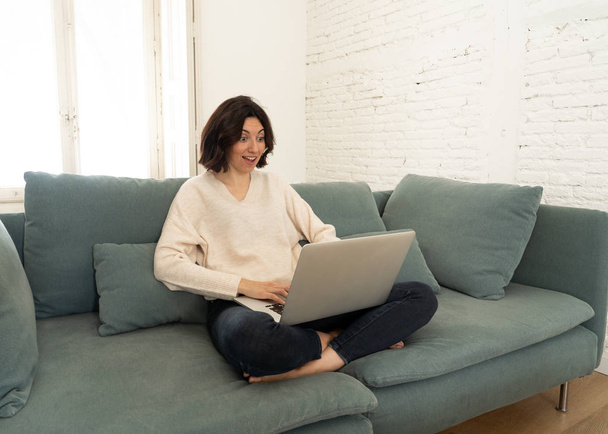 Jovencita feliz trabajando en su portátil sentada con las piernas cruzadas en el sofá. Concepto de tecnología
 - Foto, imagen