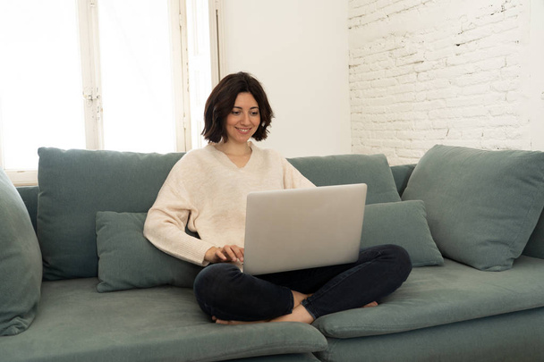 Szczęśliwa młoda kobieta pracuje na jej laptop siedzi Cross-legged na kanapie. Koncepcja technologiczna - Zdjęcie, obraz