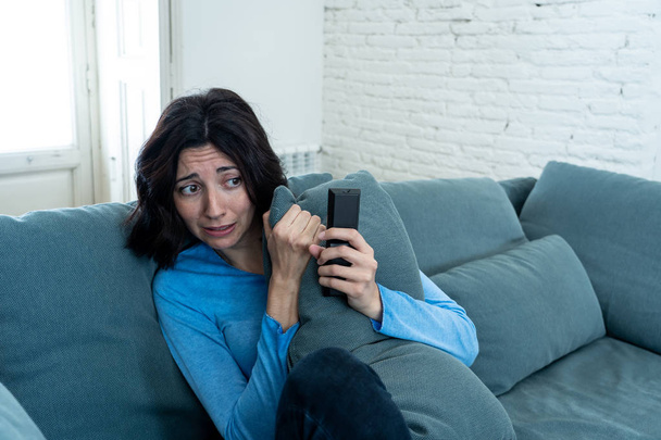 Lifestyle portrait of woman feeling scared and shocked making fear, anxiety gestures while watching horror movie on TV holding remote control. In horror and violence on TV and internet and mass media. - Фото, изображение