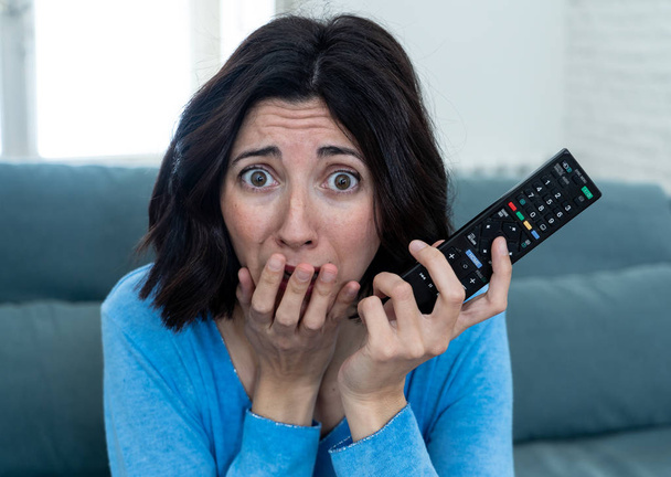 Lifestyle portrait of woman feeling scared and shocked making fear, anxiety gestures while watching horror movie on TV holding remote control. In horror and violence on TV and internet and mass media. - Foto, immagini