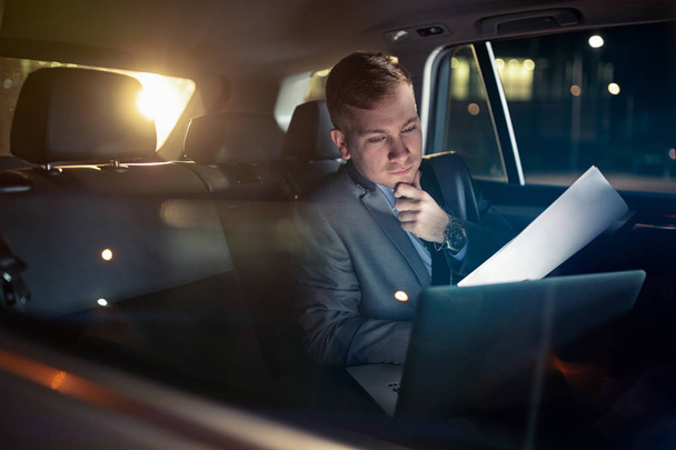 Focusing on work- businessman working on his laptop and paper on the  back seat of the car - Fotoğraf, Görsel