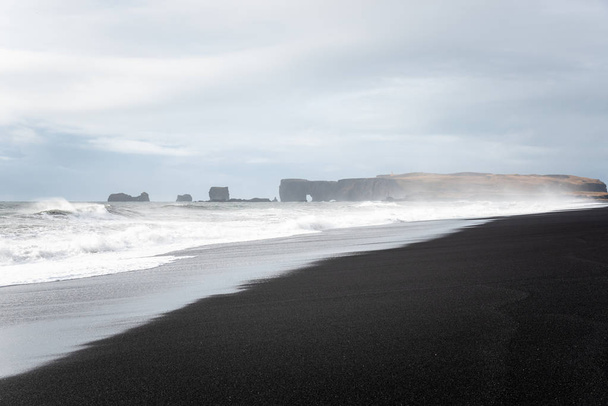Impresionante playa de arena negra desierta con acantilados a distancia parcialmente ocultos por el rocío del mar en un día nublado de otoño. Playa de Reynisfjara, Vik, Islandia
. - Foto, Imagen