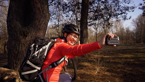 Young caucasian woman athlete tourist cyclist uses hand smart phone photo of herself selfie sitting near tree in coniferous forest outside the city. Sportswoman taking selfie with her mountain bike - Photo, Image