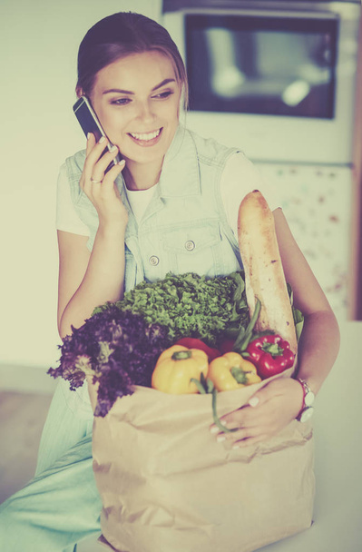 Smiling woman with mobile phone holding shopping bag in kitchen - Photo, Image
