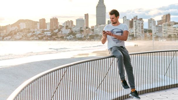 Молодий красивий чоловік зі смартфоном проти моря та панорамного вигляду міста
 - Фото, зображення
