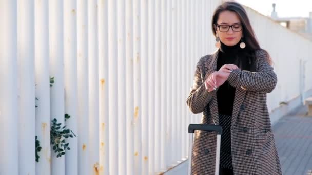 Slow motion of young busy business woman in glasses with suitcase looking at her smartwatch. Stylish successful woman with suitcase waiting for taxi. - Imágenes, Vídeo
