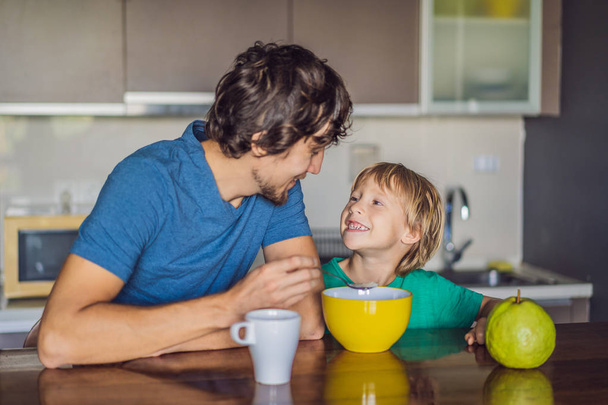 Father and son are talking and smiling while having a breakfast in kitchen - Фото, изображение