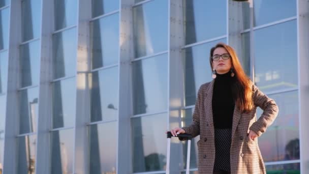 Self-confident young business woman pulling out mobile phone of her pocket and calling someone at the airport. Stylish look, elegant coat, glasses and suicase. Businesswoman waiting for the taxi. 4k video. - Πλάνα, βίντεο