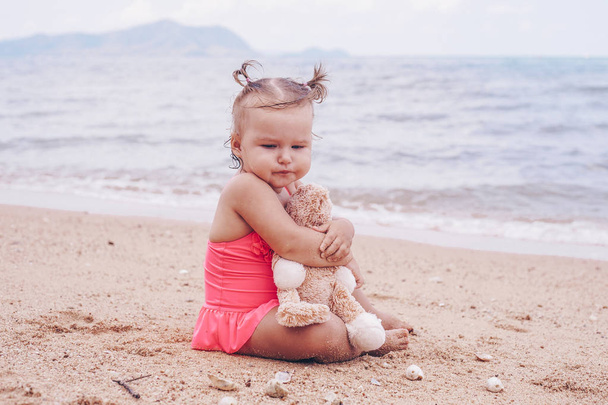 niña pensativa abrazando oso de peluche y mirando hacia otro lado mientras está sentado en la orilla del mar
. - Foto, imagen
