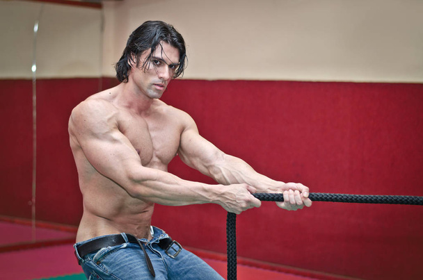 Handsome shirtless muscular man wearing jeans pulling rope indoors - Foto, afbeelding